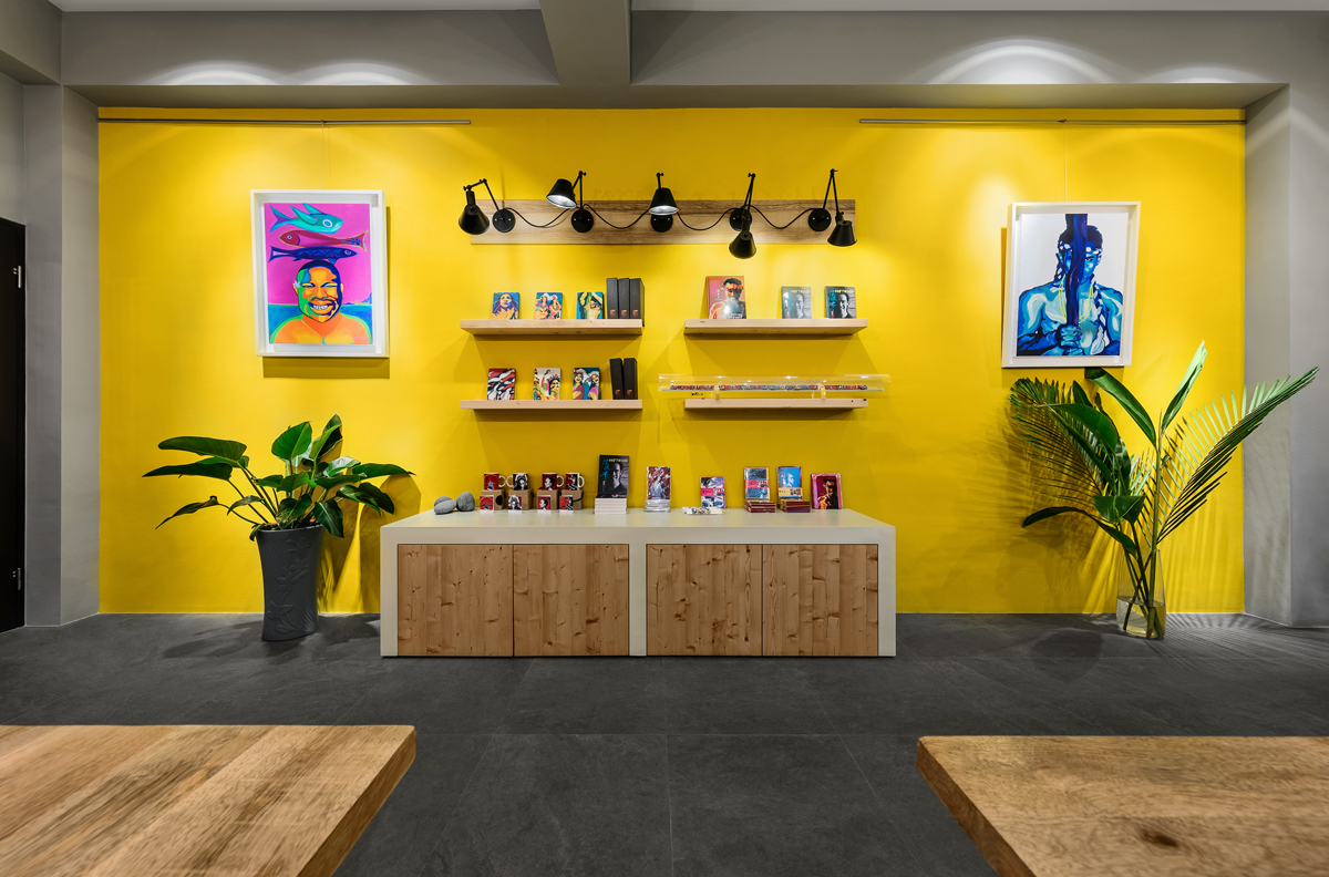 用餐區側邊展示牆面：原木色的低櫃配合大膽的黃色牆面，牆上的木質平台，陳列展示豐富的藝術作品與文藝產品。